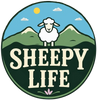 Sheepy Life Logo featuring a sheep with green mountains and a blue sky in the background with the sun and clouds, and the words "Sheepy Life" in front with a flower beneath. 